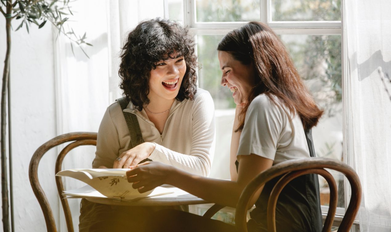 two women sitting next to a sunny bright window, looking through some papers and laughing together.