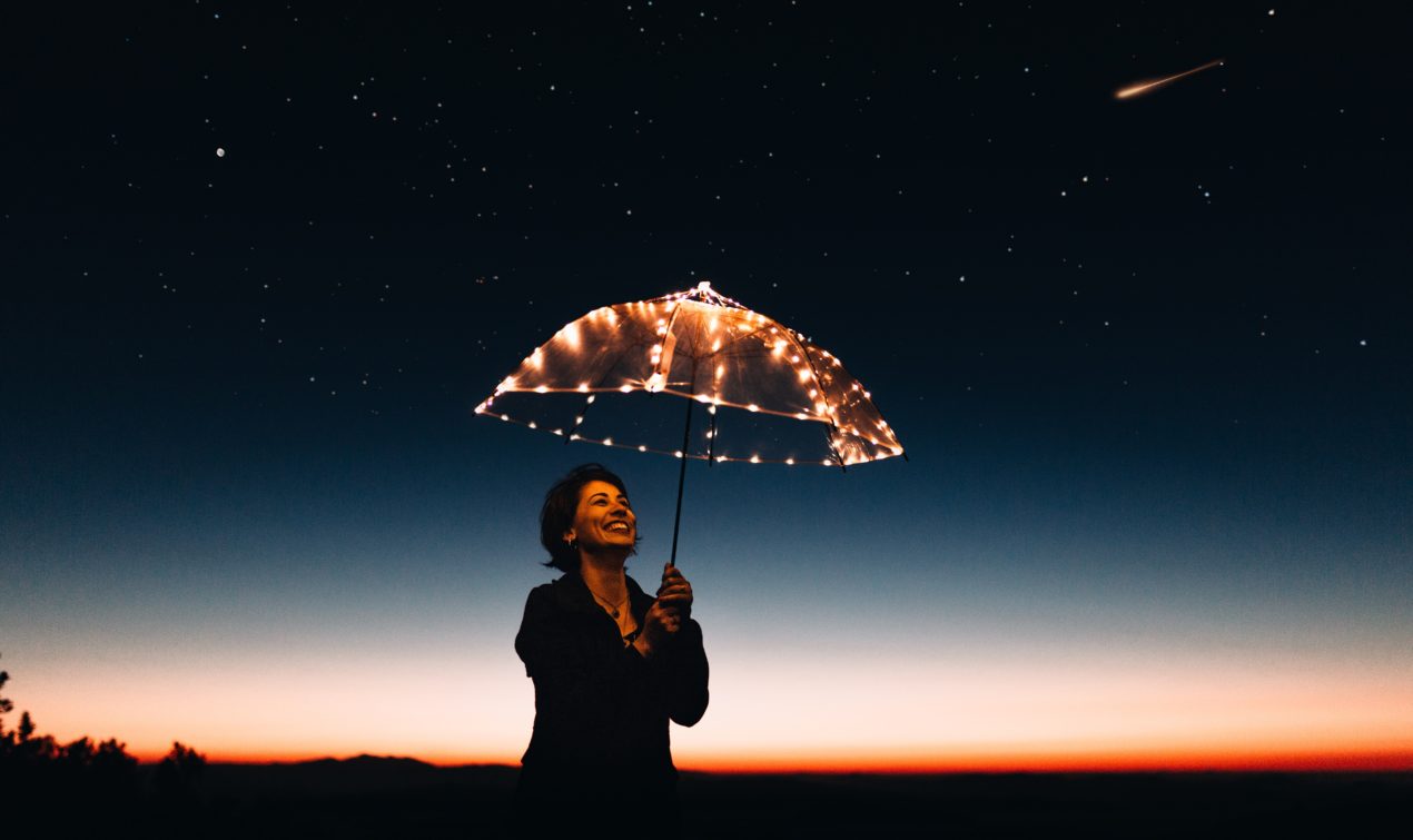 a happy, smiley woman holding a clear, lit up umbrella behind a beautiful sunset.